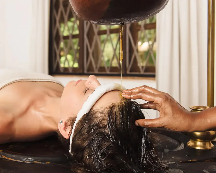A steady thread of warm oil falling from a vessel onto a guest’s forehead