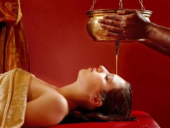 A therapist pouring a steady stream of warm medicated oil onto a guest's forehead during Shirodhara